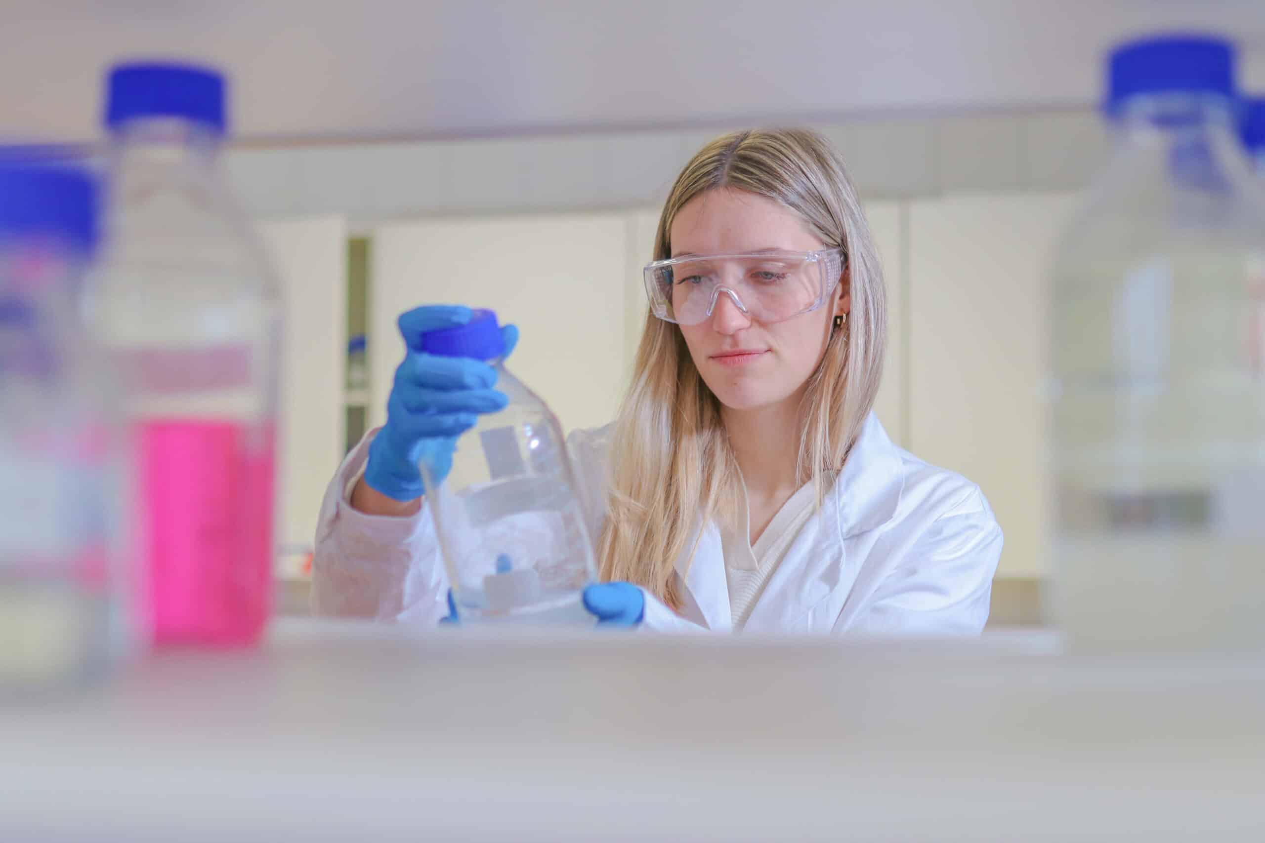 Blondine mit weißem Laborkittel, Schutzbrille und Schutzhandschuhen im Labor mit einer Glasflasche in der Hand.